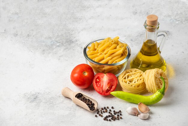 Variedad de pasta cruda, botella de aceite de oliva, granos de pimienta y verduras en el cuadro blanco.