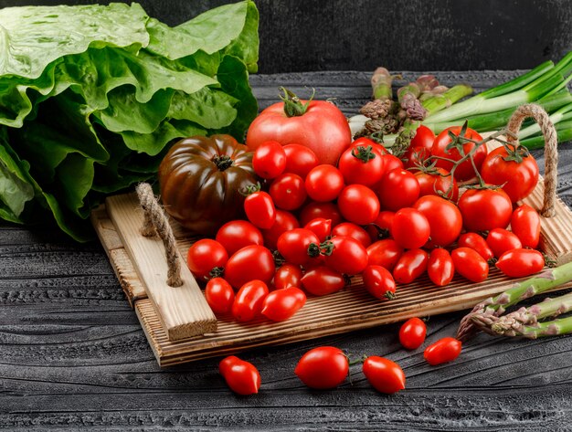 Variedad de tomates en una bandeja de madera con lechuga, espárragos, cebollas verdes, vista de ángulo alto en madera y pared oscura