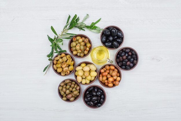 Variedad variada de aceitunas en cuencos de arcilla con hojas de olivo y una jarra de aceite de oliva vista superior en madera blanca