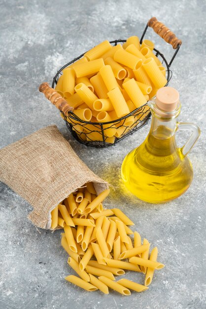 Variedades de pasta en canasta metálica y bolsa rústica con aceite de oliva sobre mesa gris.