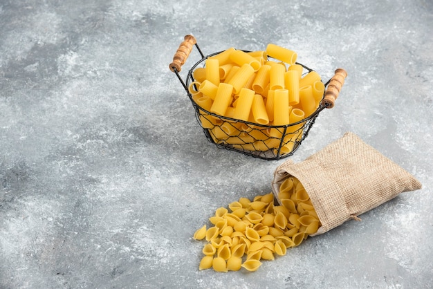 Variedades de pasta en una canasta metálica y bolsa rústica en mesa gris.