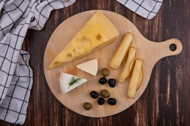 Variedades de queso de vista superior con aceitunas en un soporte con una toalla a cuadros sobre un fondo de madera