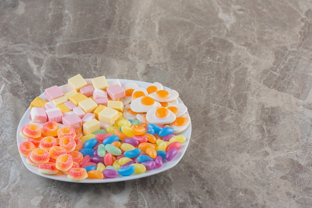 Varios tipos de caramelos de colores en un plato blanco sobre fondo gris. Caramelos de colores.