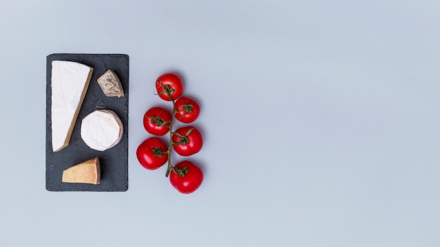 Varios tipos de queso en pizarra negra con tomates rojos sobre superficie gris con espacio de copia