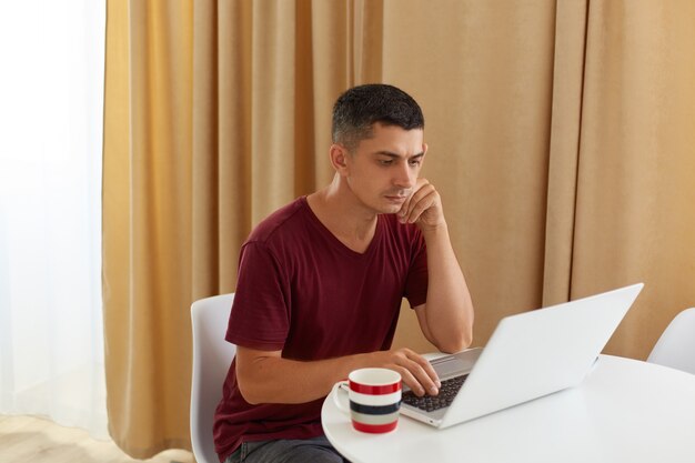 Varón adulto joven con camiseta marrón de estilo casual trabajando en línea mientras posa en la acogedora sala de estar luminosa, mirando la pantalla del portátil con expresión facial pensativa concentrada.