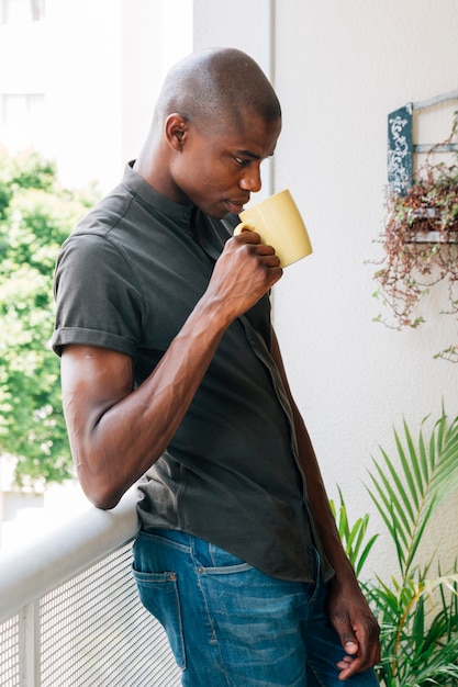 Varón africano joven que se inclina en la verja del balcón que bebe el café