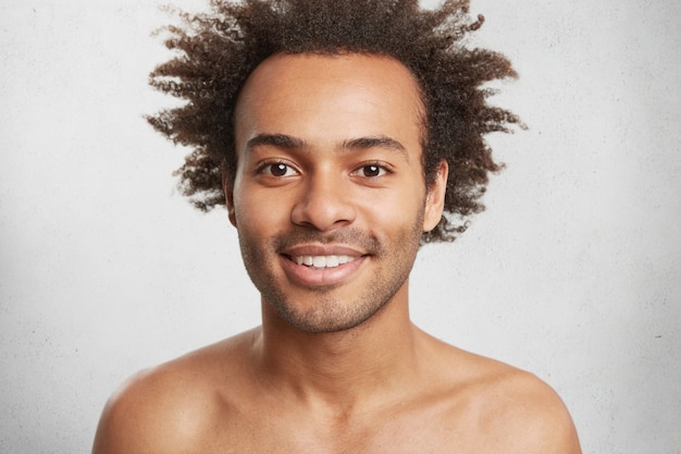 Varón afroamericano positivo desnudo con piel oscura y saludable y cabello rizado, sonríe suavemente