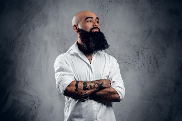 Varón árabe barbudo con tatuajes en los brazos cruzados, vestido con una camisa blanca posando sobre fondo gris.