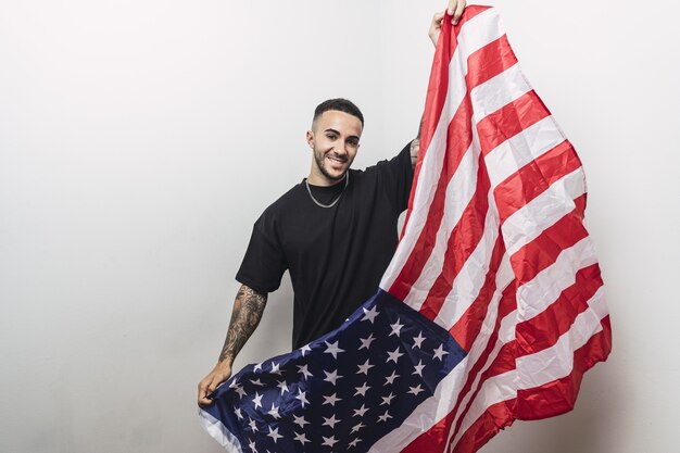 Varón español positivo con brazos tatuados posando con una bandera americana aislada en una pared blanca