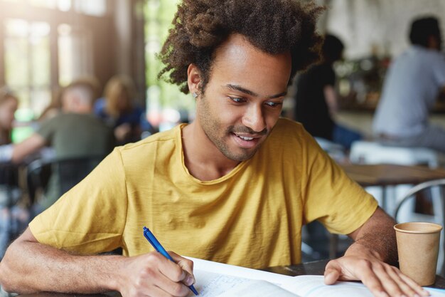 Varón estudiante enfocado con cabello oscuro y tupido y cerdas con camiseta casual escribiendo algo en su cuaderno mientras está sentado en la cantina de la universidad tomando café. Esquema de escritura de chico guapo elegante