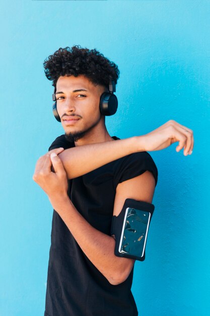 Varón étnico estirando y escuchando música con la caja del teléfono en el brazo