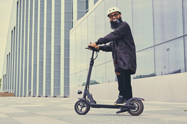 Varón hipster barbudo vapeando y montando en scooter eléctrico sobre un edificio moderno.