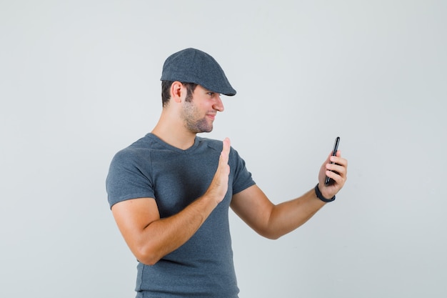 Varón joven agitando la mano en el chat de video en la gorra de la camiseta y mirando alegre