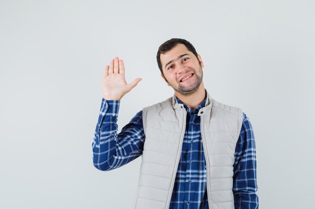 Foto gratuita varón joven agitando la mano para saludar en camisa, chaqueta sin mangas y mirando complacido, vista frontal.