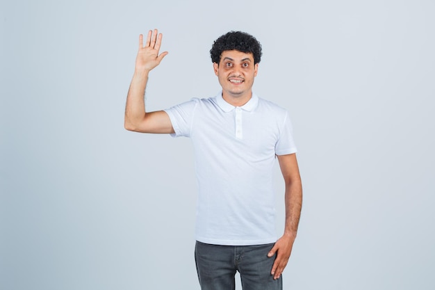 Varón joven agitando la mano para saludar en camiseta blanca, pantalones y mirando confiado, vista frontal.