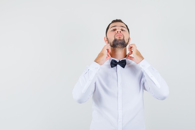 Varón joven ajustando su cuello en camisa blanca y mirando elegante, vista frontal.