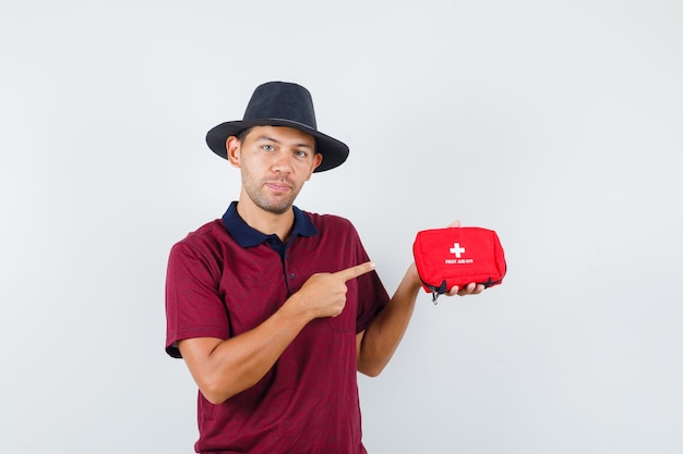 Varón joven apuntando al botiquín de ayuda rápida en camisa roja, sombrero negro y mirando serio, vista frontal.