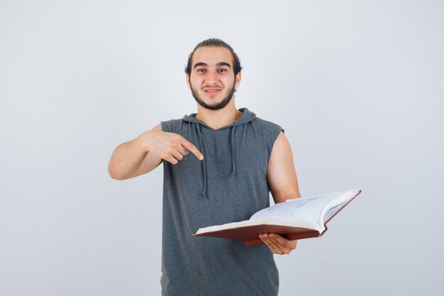 Varón joven apuntando al libro en una sudadera con capucha sin mangas y luciendo bonita, vista frontal.