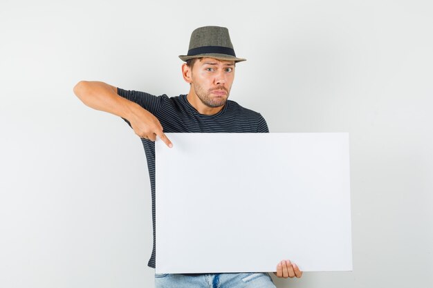 Varón joven apuntando al lienzo en blanco en jeans camiseta sombrero