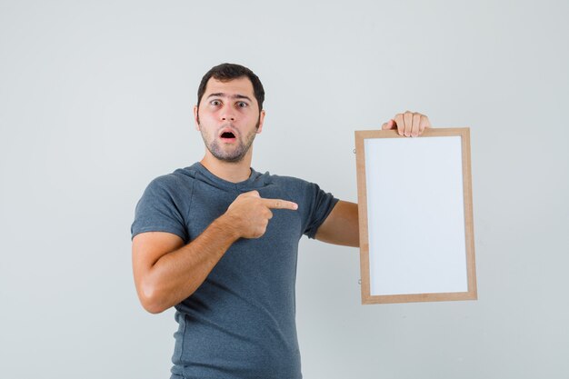 Varón joven apuntando al marco vacío en camiseta gris y mirando desconcertado