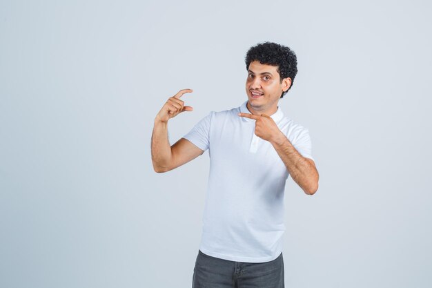 Varón joven apuntando a algo diminuto en camiseta blanca, pantalones y mirando alegre. vista frontal.