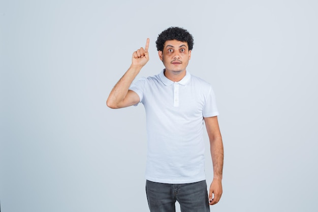 Varón joven apuntando hacia arriba con camiseta blanca, pantalón y aspecto sensato. vista frontal.