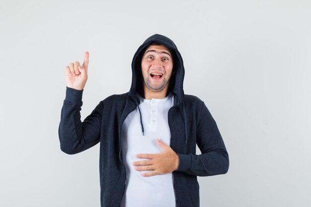 Varón joven apuntando hacia arriba en camiseta, chaqueta y mirando alegre, vista frontal.