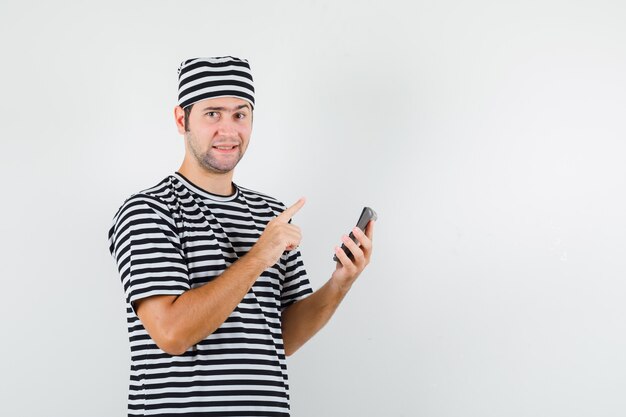 Varón joven apuntando hacia arriba, sosteniendo la calculadora en camiseta, sombrero y luciendo sensato. vista frontal.