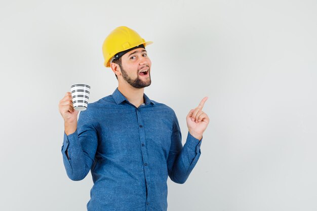 Varón joven apuntando hacia arriba, sosteniendo una taza de café en camisa, casco y mirando feliz. vista frontal.