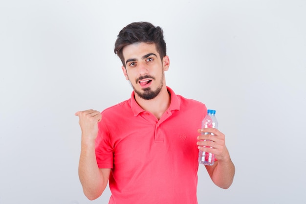 Varón joven apuntando hacia atrás con el pulgar mientras saca la lengua en la camiseta y se ve feliz, vista frontal.