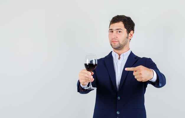 Varón joven apuntando a la bebida alcohólica en vidrio en traje, vista frontal.