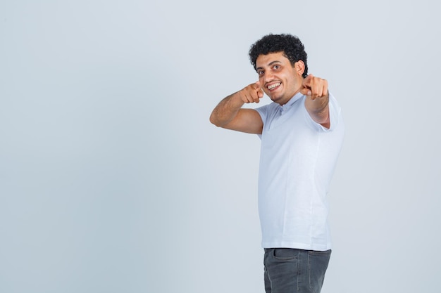 Varón joven apuntando a la cámara en camiseta blanca, pantalones y mirando feliz. vista frontal.