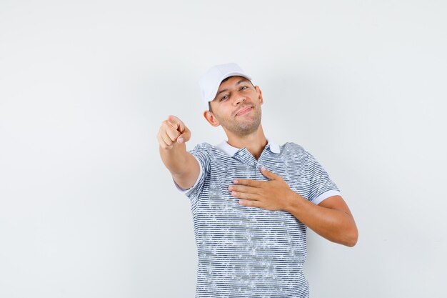 Varón joven apuntando a la cámara en camiseta y gorra y mirando alegre