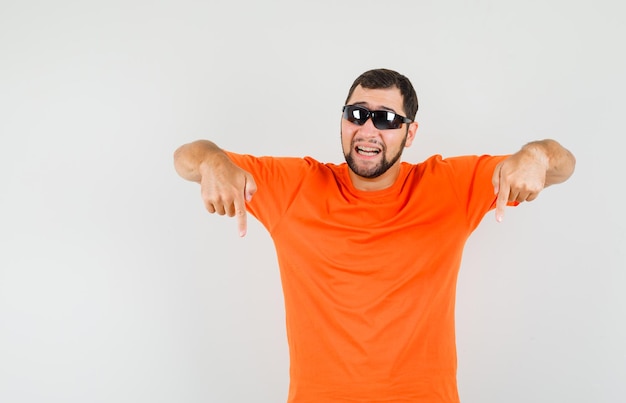 Foto gratuita varón joven apuntando con el dedo hacia abajo en camiseta naranja y mirando confiado. vista frontal.