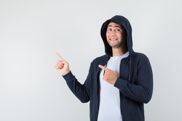 Varón joven apuntando a la esquina superior izquierda en camiseta, chaqueta y mirando alegre, vista frontal.