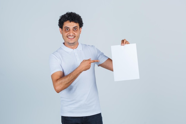 Varón joven apuntando a la hoja de papel en blanco con camiseta blanca, pantalones y mirando confiado, vista frontal.