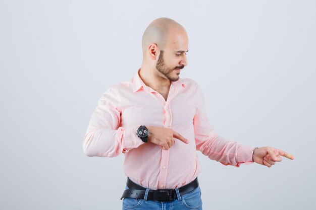 Varón joven apuntando a un lado en camisa, jeans y mirando alegre, vista frontal.