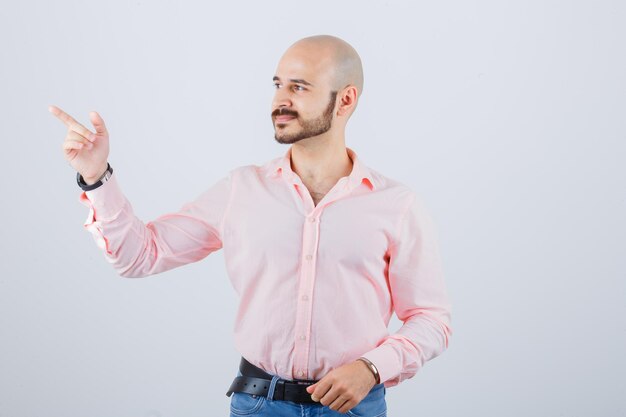 Varón joven apuntando a un lado en camisa, jeans y mirando feliz, vista frontal.