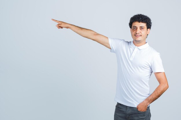 Varón joven apuntando a un lado en camiseta blanca, pantalones y mirando confiado. vista frontal.