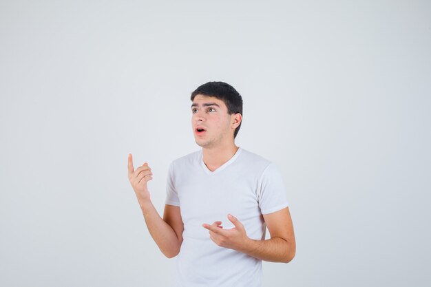 Varón joven apuntando a un lado en camiseta y mirando perplejo, vista frontal.