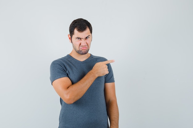 Varón joven apuntando hacia el lado derecho en camiseta gris y mirando enojado