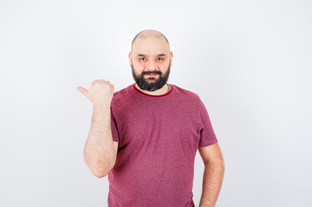 Varón joven apuntando hacia el lado derecho con el pulgar en camiseta y mirando feliz, vista frontal.