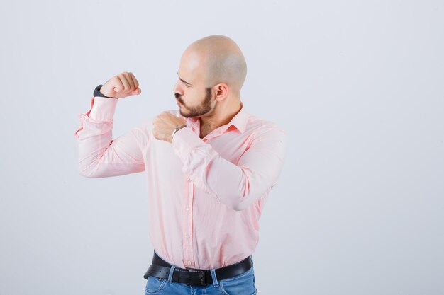 Varón joven apuntando los músculos del brazo en camisa, jeans y mirando orgulloso, vista frontal.