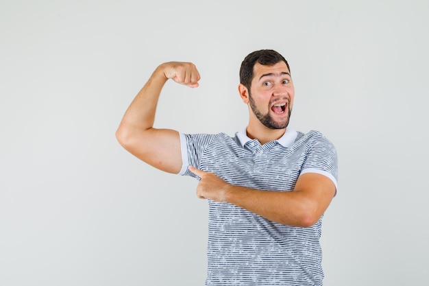 Foto gratuita varón joven apuntando a los músculos del brazo en camiseta y mirando alegre, vista frontal.