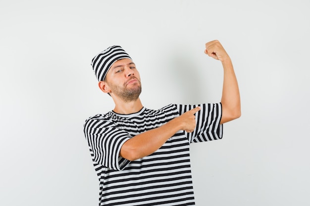 Varón joven apuntando a los músculos del brazo con sombrero de camiseta a rayas y mirando confiado