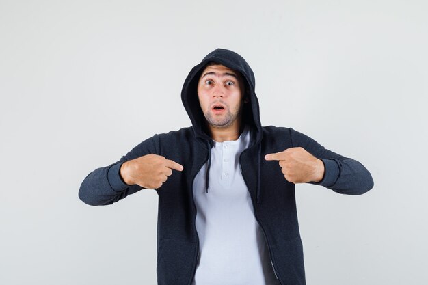 Varón joven apuntando a sí mismo en camiseta, chaqueta y mirando desconcertado. vista frontal.