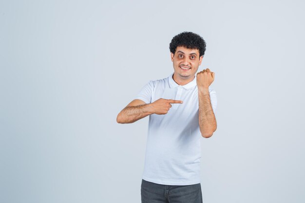 Varón joven apuntando a su brazo en camiseta blanca, pantalón y mirando alegre, vista frontal.