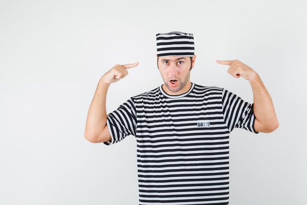 Varón joven apuntando a su cabeza en camiseta, sombrero y mirando confiado, vista frontal.