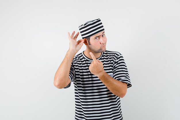 Varón joven apuntando a su oreja en camiseta, sombrero y mirando pensativo. vista frontal.