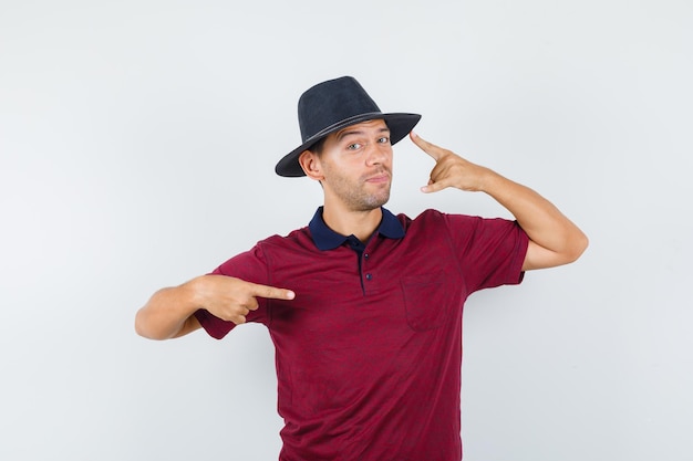 Foto gratuita varón joven apuntando a su sombrero y al lado en camiseta, sombrero y mirando confiado. vista frontal.